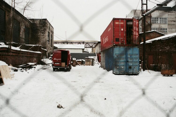 Snow-covered industrial yard with shipping containers, behind chain link fence.